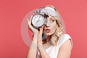 Portrait of charming young woman 20s wearing sleeping mask holding alarm clock after awakening