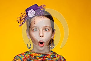 Portrait of a cheerful 8-year-old girl in a miniature hat and in a multi-colored sweater. Yellow background