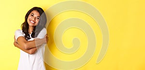 Portrait of cheerful african american woman, hugging herself and smiling pleased, standing over yellow background