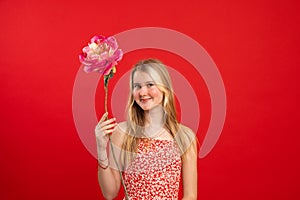 Portrait of cheerful amazing teenage girl with fair hair holding raising hand with flower pink peony on red background.