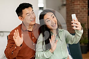 Portrait of cheerful asian married spouses using cellphone video calling and waving hello to phone webcamera