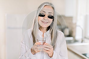 Portrait of cheerful attractive elderly woman model with black antiwrinkle eye patches, smiling to camera, while photo