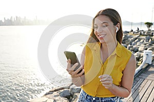 Portrait of cheerful attractive young woman wearing earphones using mobile phone outdoor with sunset in background. People and