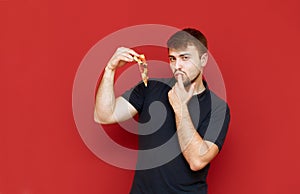 Portrait of cheerful bearded man with tasty slice of pizza in hands on red background, looks into camera, smiles and licks finger