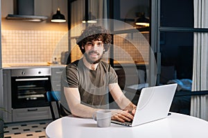 Portrait of cheerful bearded young man using typing on laptop sitting at table in kitchen room with modern interior