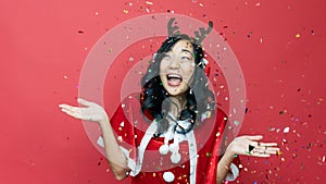 Portrait of a cheerful beautiful girl wearing red santy costume standing under confetti rain and celebrating  over red