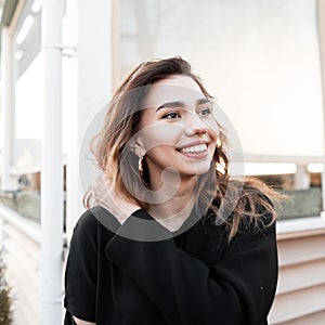 Portrait of a cheerful beautiful young woman with an attractive smile in a fashionable black coat near a wooden summer open cafe