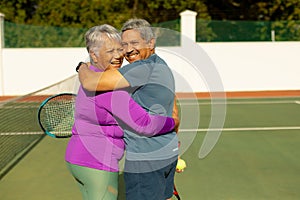 Portrait of cheerful biracial senior couple embracing while standing at tennis court on sunny day