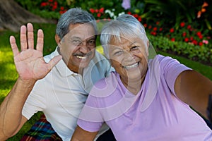 Portrait of cheerful biracial senior man waving hand while enjoying picnic with senior wife in park