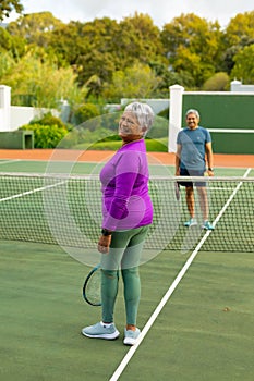 Portrait of cheerful biracial senior woman playing tennis with husband at tennis court against trees