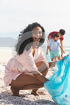 Portrait of cheerful biracial woman collecting waste in blue plastic bag with family at beach