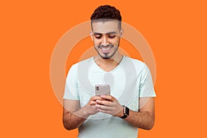 Portrait of cheerful brunette man using cellphone and smiling, reading good news. indoor studio shot isolated on orange background