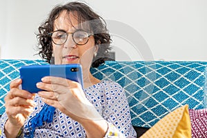 Cheerful brunette senior woman using smartphone while sitting on sofa