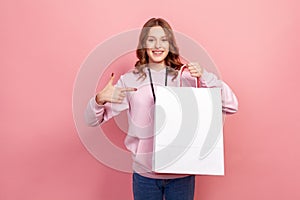 Portrait of cheerful brunette teenage girl in hoodie pointing finger at paper bags in hand, looking with toothy smile, shopping