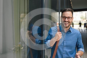 Portrait of cheerful businessman carrying laptop bag standing by window in the city