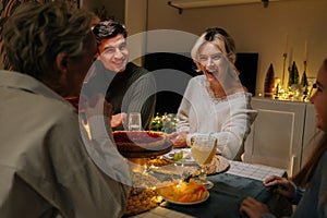 Portrait of cheerful Caucasian family celebrating Christmas, chatting at dinner holiday table in dark living room with