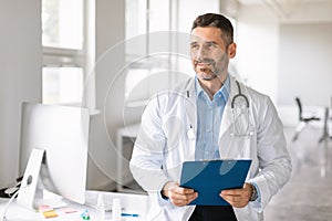 Portrait of cheerful confident male therapist posing with clipboard in hands, standing near his workplace in clinic