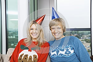 Portrait of cheerful couple wearing Christmas sweaters and party hats in living room at home