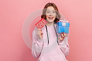 Portrait of cheerful curly haired teenage girl in hoodie holding sale word and wrapped gift box, low price on presents, discount