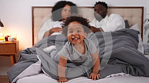 Portrait of cheerful cute little African American boy sitting on bed with parents talking at background. Joyful charming