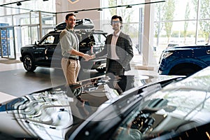 Portrait of cheerful dealer wearing business suit and handsome customer male handshaking in front of automobiles in car