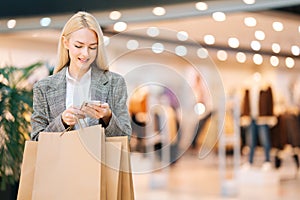 Portrait of cheerful elegance blonde young woman in stylish clothes using mobile phone holding shopping paper bags with