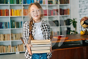 Portrait of cheerful elementary child school girl holding stack of books in library at school, looking at camera.
