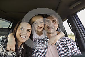 Portrait of cheerful family with kid on road trip. Girl hugging her young parents inside of car during summer vacation