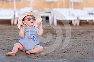 Portrait of cheerful fashionable baby girl sitting on beach coastline and enjoying summer vacation