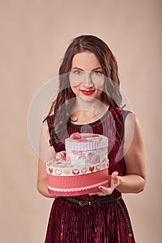 Portrait of cheerful girl dressed in dress hol.ding gift box and looking at camera, on beige background. Postcard template for