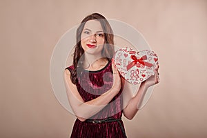 Portrait of cheerful girl dressed in dress hol.ding gift box and looking at camera, on beige background. Postcard template for