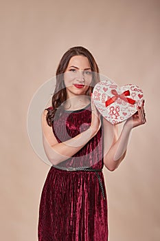 Portrait of cheerful girl dressed in dress hol.ding gift box and looking at camera, on beige background. Postcard template for