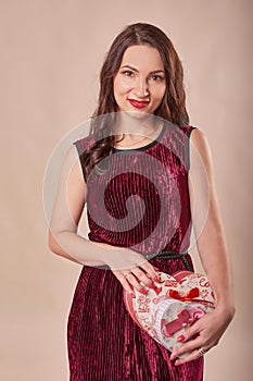 Portrait of cheerful girl dressed in dress hol.ding gift box and looking at camera, on beige background. Postcard template for