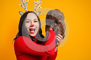 Portrait of a cheerful girl in a red sweater and with deer horns lifts up a cute cat