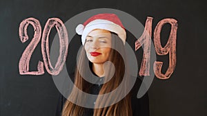Portrait of a cheerful girl in Santa`s hat, against a black slate wall. Happy New Year 2019 Concept
