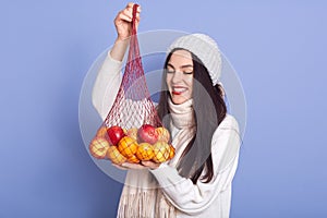 Portrait of cheerful good looking brunette looking at mandarines and apples, holding net handbag in hands, being in good mood,