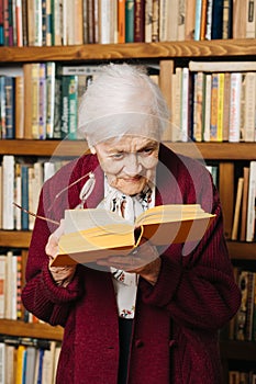 Portrait of cheerful grey haired woman reading book near bookshelf at home