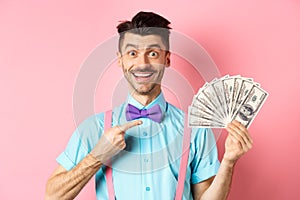 Portrait of cheerful guy with moustache and bow-tie pointing at money, holding dollars and smiling excited, standing