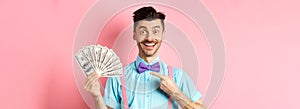 Portrait of cheerful guy with moustache and bow-tie pointing at money, holding dollars and smiling excited, standing