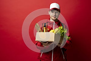 Portrait of cheerful handsome young delivery man holding grocery shopping box isolated on red background. Online