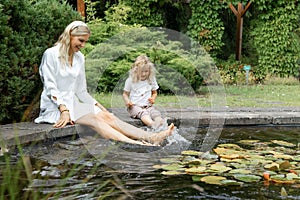 Portrait of cheerful family of young woman and little girl sitting splashing with bare feet in water on edge of pond.