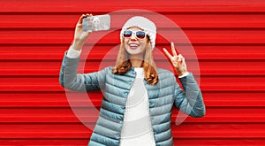 Portrait cheerful happy laughing young woman taking selfie by smartphone wearing a white hat, jacket on red background