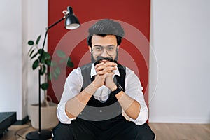 Portrait of cheerful Indian businessman wearing elegant formalwear sitting on chair and smiling looking at camera. Front