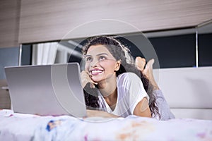 Cheerful woman daydreaming with laptop on bed