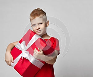 Portrait of cheerful kid boy in red t-shirt holding big present box with ribbon in hands, carrying it