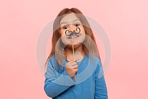 Portrait of cheerful little girl holding paper mustache and smiling to camera, carefree child playing, making funny face