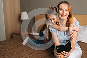 Portrait of cheerful little girl hugging to beautiful mother holding mobile phone and looking at camera, chatting on