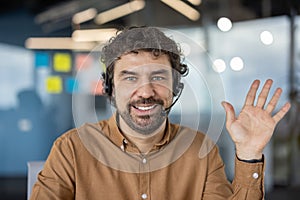 Portrait of a cheerful male customer service representative wearing a headset and waving in a modern office setting.