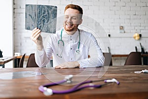 Portrait of cheerful male doctor in white uniform examining brain computerized tomography scan sitting at desk in