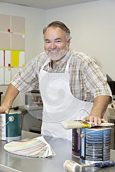 Portrait of a cheerful mature store clerk with paint cans and brush in hardware shop